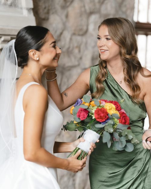 Lindsay in a green dress touching up a smiling bride holding a bright bouquet
