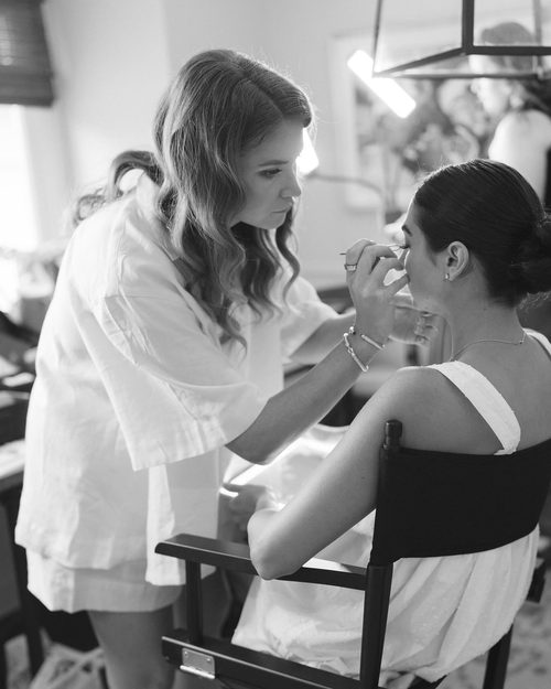 Black and white photo of Lindsay applying lashes to a client seated in a director's chair